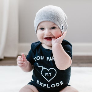 May include: A baby wearing a black bodysuit with the text "You make my heart explode" and a grey beanie. The baby is sitting on a white rug and smiling.