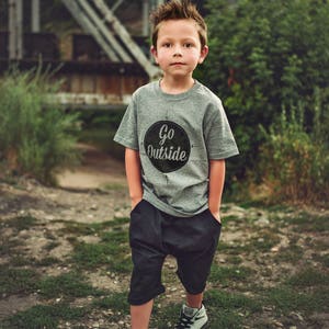 Pode incluir: Um menino pequeno vestindo uma camiseta cinza com um c&iacute;rculo preto e o texto "Go Outside" impresso nela. Ele est&aacute; vestindo shorts pretos e t&ecirc;nis cinza.