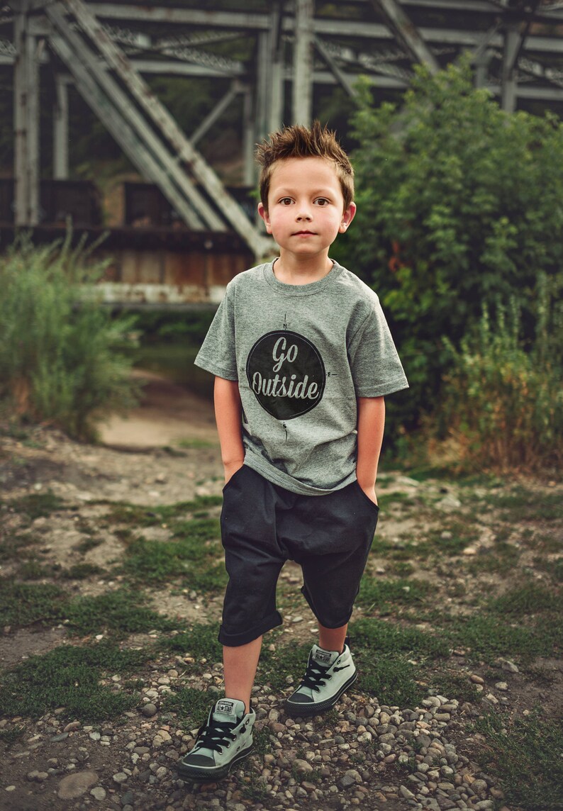 Pode incluir: Um menino pequeno vestindo uma camiseta cinza com um c&iacute;rculo preto e o texto "Go Outside" impresso nela. Ele est&aacute; vestindo shorts pretos e t&ecirc;nis cinza.
