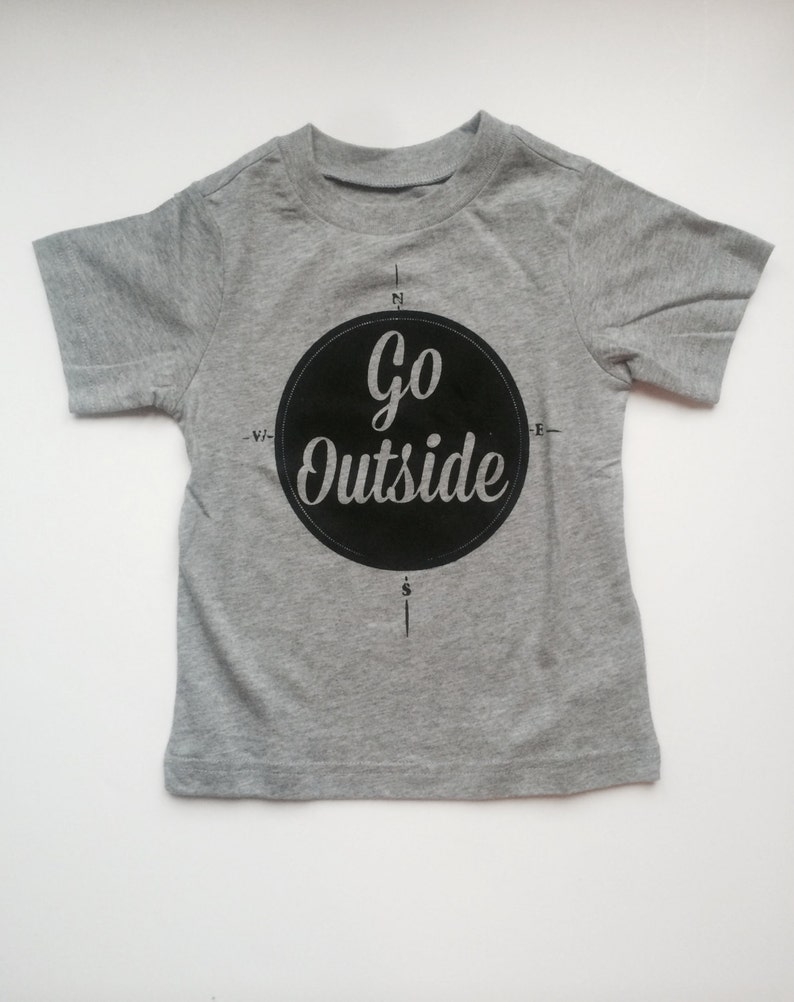 Pode incluir: Uma camiseta cinza de manga curta com um gr&aacute;fico circular preto que diz "Go Outside" e um design de b&uacute;ssola.
