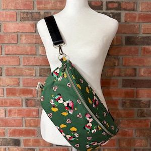 May include: A green crossbody bag featuring a repeating pattern of Mickey and Minnie Mouse dressed as elves, interspersed with candy canes. The bag has a gold-tone zipper closure, a black adjustable strap with a metal clip, and a small white fabric tag.