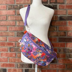May include: A crossbody fanny pack featuring a vibrant purple, orange, and pink tie-dye background with a repeating pattern of small purple dragon characters. The bag includes a purple adjustable strap, a top zipper closure, and a small orange tab.