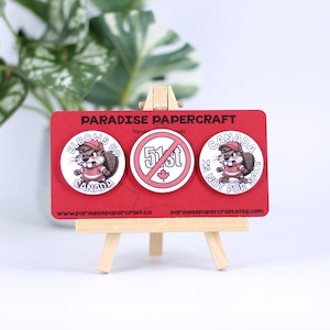 Puede incluir: Tres botones redondos con fondo rojo y texto blanco. El primer botón presenta un castor de dibujos animados con un jersey de hockey y el texto "Elbows Up Canada". El segundo botón presenta un círculo rojo con una línea diagonal que lo atraviesa y el texto "51st". El tercer botón presenta un castor de dibujos animados con un jersey de hockey y el texto "Canada Is Not For Sale".