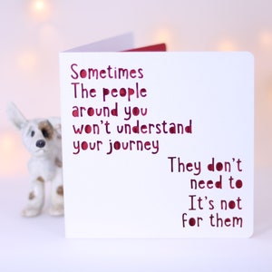 May include: A white greeting card with red cut-out text that reads: "Sometimes the people around you won't understand your journey. They don't need to. It's not for them." A small ceramic dog figurine is in the background.