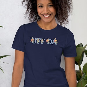 Peut inclure: Un t-shirt bleu marine avec le texte "UFF DA" en lettres bleu clair avec des accents floraux.
