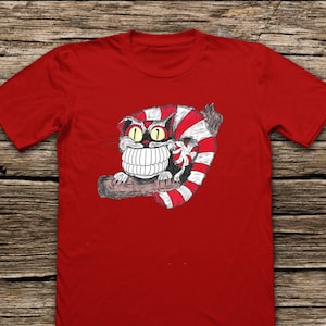 May include: Red t-shirt with a black and white illustration of a Cheshire Cat with a wide grin, wearing a red and white striped hat.