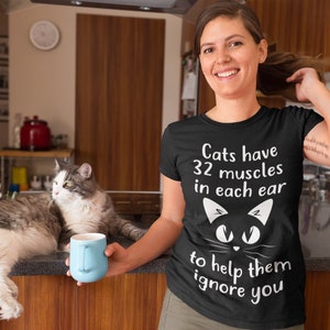 May include: A black t-shirt with white text that reads "Cats have 32 muscles in each ear to help them ignore you". A black cat face with white eyes is above the text. A woman is wearing the shirt and holding a blue mug. A cat is sitting on a counter behind her.