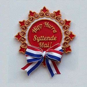 May include: A red and gold decorative ornament with a white ribbon bow. The ornament features the text "Hipp Hurra Syttende Mai!" in gold lettering.