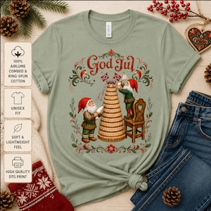 以下が含まれることがあります： セージグリーンの半袖Tシャツ。2体のノームが背の高い段重ねのケーキを飾っているグラフィックが描かれています。赤い帽子と緑の服を着たノームの上には「God Jul」の文字がプリントされています。デザインには2本の小さなノルウェー国旗が含まれています。