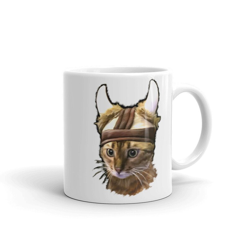 May include: White ceramic coffee mug with a brown tabby cat wearing a brown leather helmet with horns. The cat has green eyes and is looking to the left.