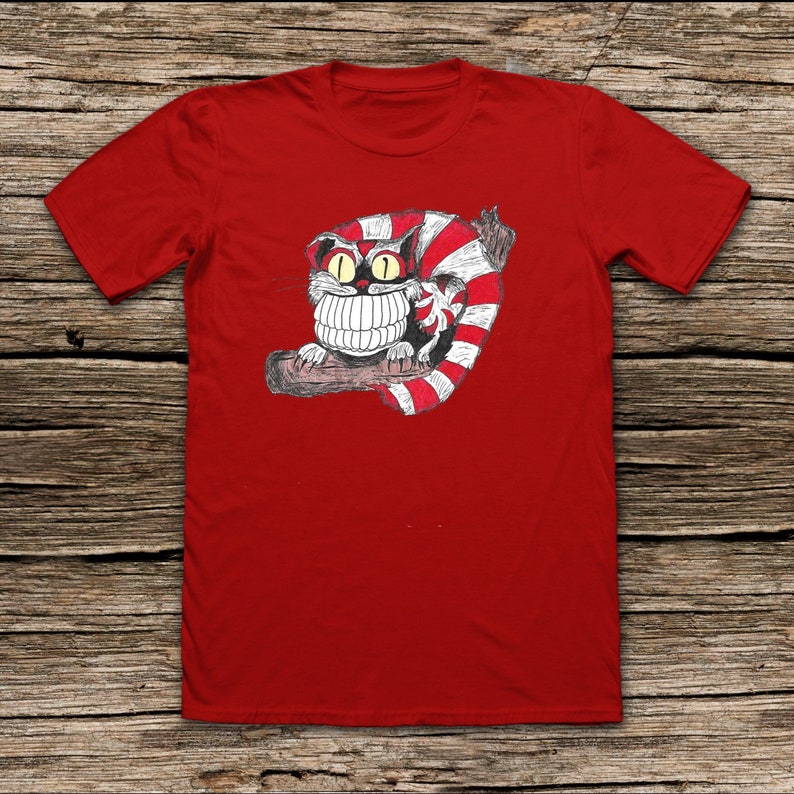 May include: Red t-shirt with a black and white illustration of a Cheshire Cat with a wide grin, wearing a red and white striped hat.