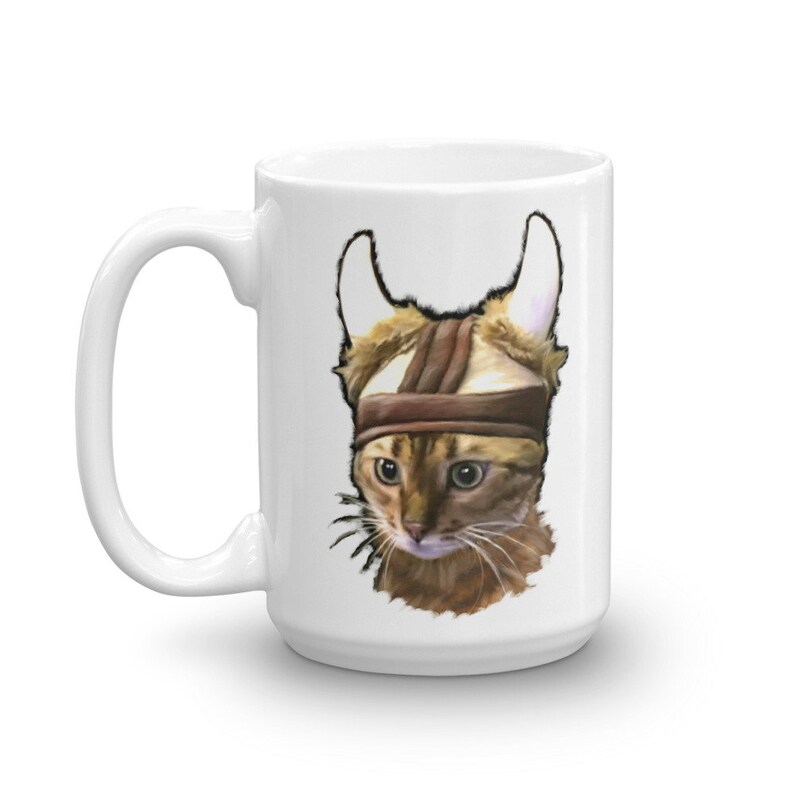May include: White ceramic coffee mug with a brown tabby cat wearing a brown leather helmet with horns. The cat has green eyes and a serious expression.