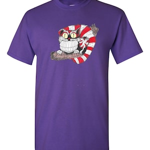 May include: Purple t-shirt with a cartoon Cheshire Cat from Alice in Wonderland. The cat is red and white with a large grin and big eyes.