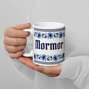 Könnte beinhalten: Weiße Keramiktasse mit dem Wort "Mormor" in dunkelblauer gotischer Schrift. Die Tasse hat ein blaues Blumendesign mit einem dunkelblauen Rand. Die Tasse wird von einer Person gehalten.