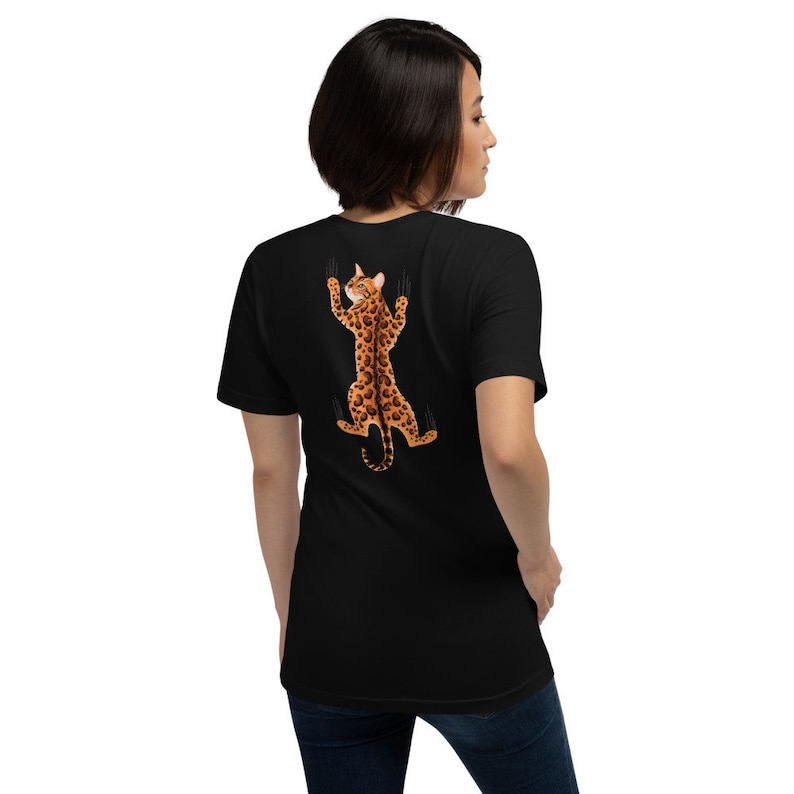 Pode incluir: Camiseta preta com um gr&aacute;fico de um gato marrom e preto manchado agarrado &agrave; parte de tr&aacute;s da camiseta com as garras estendidas.