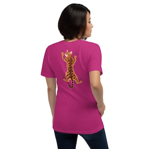 Pode incluir: Uma camiseta rosa com um gr&aacute;fico de um gato com estampa de leopardo escalando uma parede. O gato est&aacute; de costas para o espectador e tem as garras estendidas.