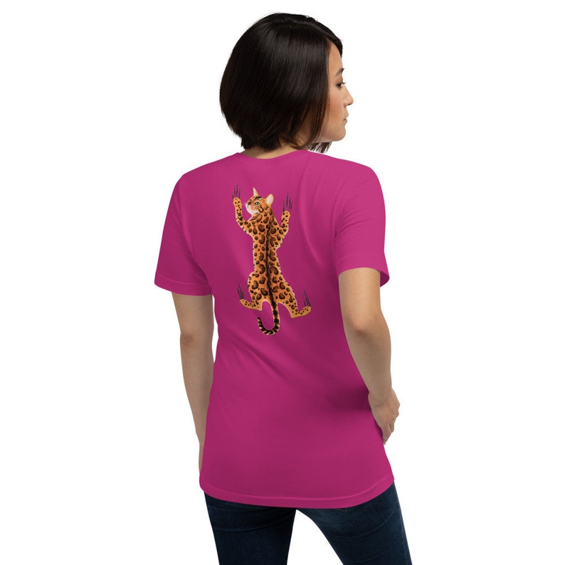 Pode incluir: Uma camiseta rosa com um gr&aacute;fico de um gato com estampa de leopardo escalando uma parede. O gato est&aacute; de costas para o espectador e tem as garras estendidas.