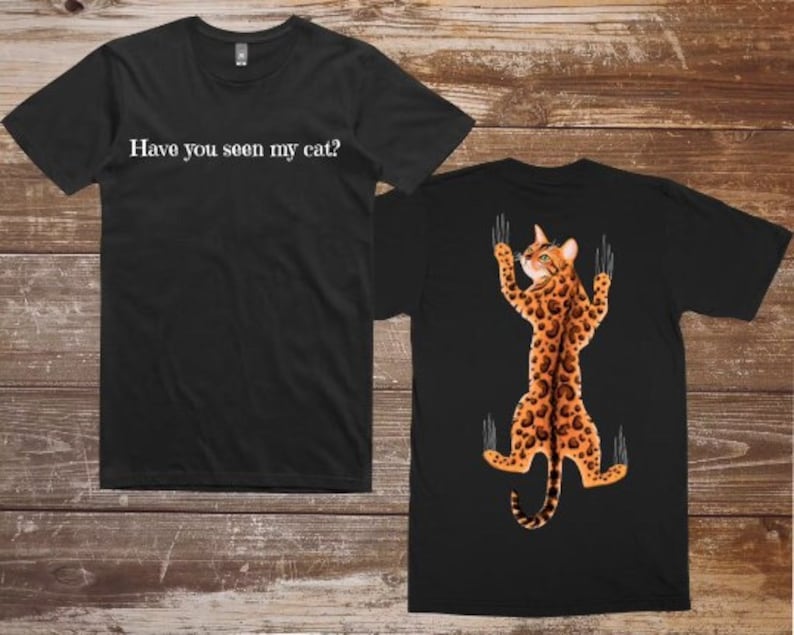 Pode incluir: Camiseta preta com o texto "Voc&ecirc; viu meu gato?" na frente e um gato preto com manchas laranja agarrado &agrave; parte de tr&aacute;s da camiseta.