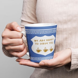 May include: A white-handled coffee mug with a blue background and a white shield design. The shield has the text "WE ARE HAPPY TO SERVE YOU" and three coffee cup illustrations. The mug has a Greek key border.