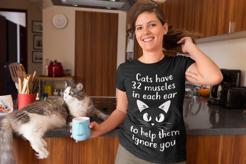May include: A black t-shirt with white text that reads "Cats have 32 muscles in each ear to help them ignore you". A black cat face with white eyes is above the text. A woman is wearing the shirt and holding a blue mug. A cat is sitting on a counter behind her.