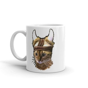 May include: White ceramic mug with a brown tabby cat wearing a brown leather helmet with horns. The cat is looking to the left of the image.
