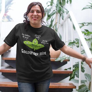 A woman stands on stairs wearing a black t-shirt with a graphic of mint leaves and the words Encourage mint.