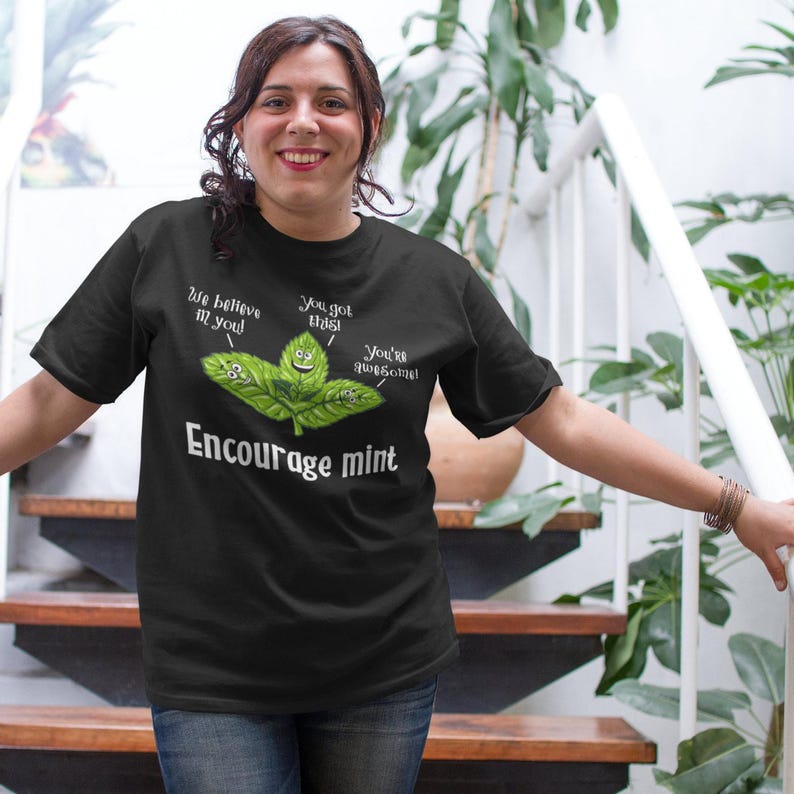 A woman stands on stairs wearing a black t-shirt with a graphic of mint leaves and the words Encourage mint.
