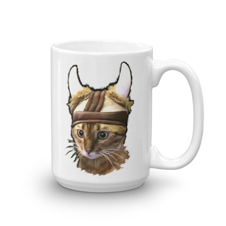 May include: White ceramic coffee mug with a brown tabby cat wearing a brown leather viking helmet. The cat is looking to the left of the image.