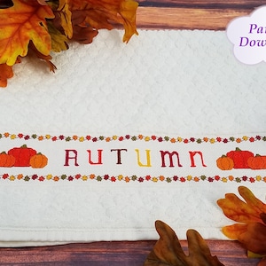 Puede incluir: Una toalla de cocina blanca con un diseño de otoño bordado. El diseño presenta la palabra "AUTUMN" en letras rojas, naranjas y amarillas, con tres calabazas a cada lado de la palabra. Las calabazas son naranjas y marrones con tallos verdes. El diseño está rodeado por un borde de pequeñas hojas de colores.
