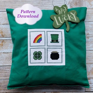 May include: Green pillow with a white square in the center. The square is divided into four smaller squares, each containing a different St. Patrick's Day themed cross-stitch design: a rainbow, a leprechaun hat, a four-leaf clover, and a pot of gold. The pillow has the text "Pattern Download" in a purple cloud above the square. A green shamrock sign with the text "Happy Go Lucky" is hanging above the pillow.