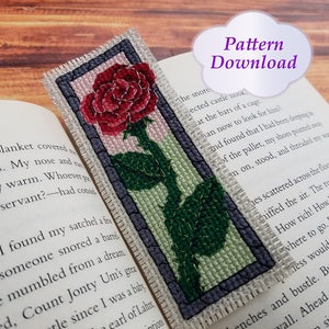May include: A cross-stitch bookmark featuring a red rose with green leaves and a light pink background. The bookmark is framed with a dark gray border and a white trim.