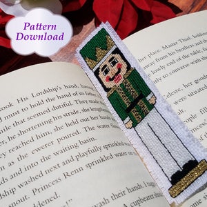 May include: A cross-stitch bookmark featuring a Nutcracker soldier. The bookmark is white with a green, gold, and white Nutcracker design. The bookmark is on a book with text. The text on the book reads "Pattern Download".