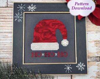 Op de afbeelding: Een ingelijste kerstkruissteek met een rode kerstmuts met een witte rand en een sneeuwvlokaccent. De muts heeft een wervelend patroon en de woorden "HO HO HO" zijn eronder geborduurd. De lijst is donkergrijs met witte sneeuwvlokaccenten en de tekst "Pattern Download".