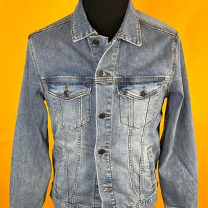 May include: A light blue denim jacket with a classic collar, button-down front, and two chest pockets. The jacket is displayed on a black mannequin against a yellow background. The jacket has dark stitching and a tailored fit.