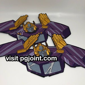 May include: Two purple and gold embroidered iron-on patches featuring a robot character with the text "visit pgjoint.com".