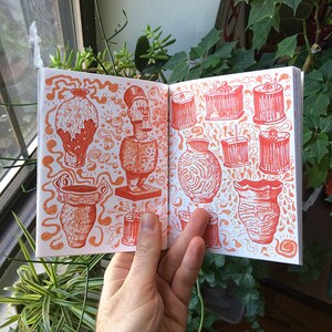 May include: Open sketchbook with orange ink illustrations of various vases, statues, and cylindrical objects. The drawings are detailed and set against a backdrop of swirling patterns. The sketchbook is held open by a hand, showcasing the artwork.
