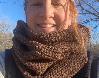 Könnte beinhalten: Ein brauner gehäkelter Cowl-Schal. Der Schal ist aus einem strukturierten Garn mit Farbtupfern gefertigt. Der Cowl wird um den Hals getragen und ist ein warmes Accessoire. Der Hintergrund ist ein klarer blauer Himmel und kahle Äste.