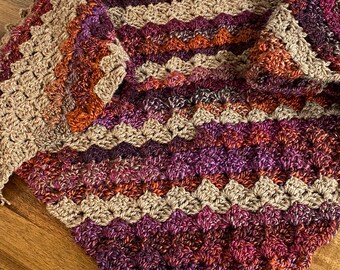 Könnte beinhalten: Ein handgehäkelter Schal in einer warmen Farbpalette aus Beige, Orange, Burgunderrot und Lila. Der Schal hat ein strukturiertes, muschelartiges Stichmuster und einen gewellten Rand. Der Artikel liegt auf einer Holzoberfläche.