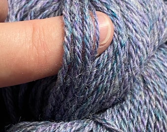 Könnte beinhalten: Nahaufnahme eines Garnknäuels in den Farben Lila, Blau und Grau. Das Garn ist weich und flauschig mit einem dezenten Glanz.
