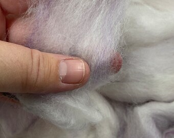 Könnte beinhalten: Nahaufnahme einer Handvoll weißer Wollroving mit dezenten lilafarbenen Streifen. Die weiche, flauschige Textur ist sichtbar, was auf ein Bastelmaterial zum Filzen oder Spinnen hindeutet. Die Fasern werden in einer Hand gehalten.