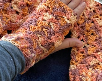 Könnte beinhalten: Gehäkelte Fingerhandschuhe und ein passender Schal in Orange-, Pfirsich- und Brauntönen. Das Lochmuster ist deutlich zu sehen und unterstreicht die handwerkliche Qualität des Sets. Die Handschuhe werden an einer Hand gezeigt, der Schal ist teilweise sichtbar.