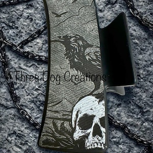 May include: Black hair clip with a raven and skull design. The raven is perched on a branch with a mountain range in the background. The clip has the text "Three Dog Creations".
