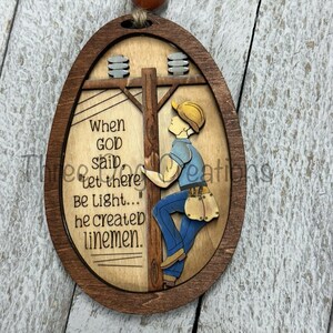 May include: A wooden oval ornament with a cartoon image of a lineman climbing a utility pole. The ornament has the text "When GOD said "Let there be light..." he created linemen."