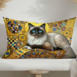 Op de afbeelding: Een decoratief kussen met een witte en grijze kat met blauwe ogen, liggend op een gouden achtergrond met geometrische patronen. De kat wordt omringd door cirkels, vierkanten en andere vormen in tinten geel, blauw en groen.