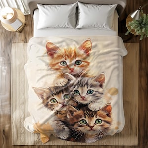 May include: A cream-colored blanket featuring a stack of four adorable kittens. The kittens have orange, gray, and brown fur with green and blue eyes. The blanket is on a bed with white pillows.