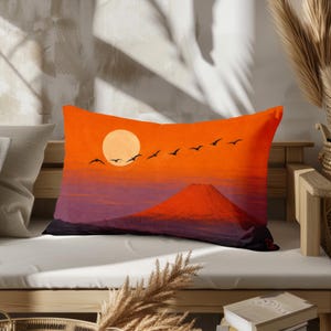 Könnte beinhalten: Ein rechteckiges Dekokissen mit einer lebendigen Sonnenuntergangsszene, einem leuchtend orangefarbenen Himmel, einer großen blassen Sonne und der Silhouette eines Berggipfels. Ein Vogelschwarm fliegt über den Horizont, mit dunklen Violett- und Schwarztönen an der Basis.