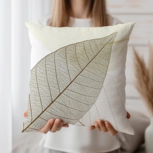 Op de afbeelding: Wit decoratief kussen met een groot, gedetailleerd bladerontwerp in bruin- en beigetinten. Het kussen heeft een zacht, neutraal kleurenpalet en een natuurlijk, botanisch thema. De ingewikkelde nerven van het blad zijn duidelijk zichtbaar.