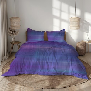 Op de afbeelding: Een bed met een dekbedovertrek en twee kussenslopen met een paars, blauw en roze gestreept patroon. Het beddengoed heeft een horizontaal streepontwerp. De kamer heeft een neutrale kleurstelling met geweven armaturen.
