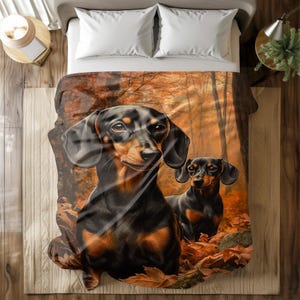 Könnte beinhalten: Eine Decke mit einer digitalen Illustration von zwei Dackel-Hunden in einer Waldszene. Die Hunde sind schwarz und braun, vor dem Hintergrund von orangefarbenen und braunen Herbstblättern und Bäumen. Die Decke liegt auf einem Bett mit weißen Kissen.