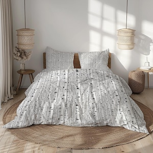 Op de afbeelding: Een wit en grijs beddengoed met berkenpatroon. De set bevat een dekbedovertrek en twee kussenslopen. Het ontwerp toont verticale lijnen die lijken op berkenstammen. Het beddengoed ligt op een bed met een houten hoofdbord en een rond, geweven tapijt.